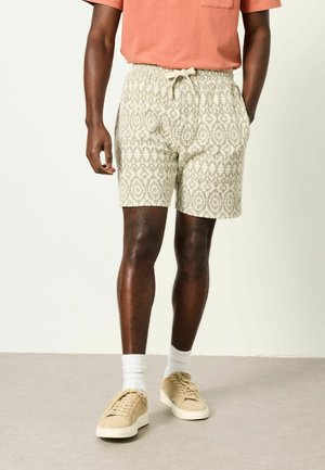 Man wearing beige patterned shorts with drawstring, white socks, beige sneakers, and a salmon-colored short sleeve shirt.