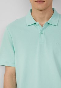 Polo mint vert en tissu texturé, avec un col classique, une patte de boutonnage à deux boutons et un discret logo sur la poitrine.