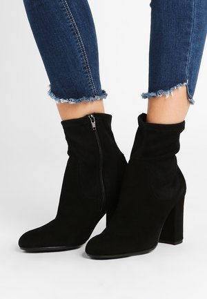 Bottines en daim noir avec fermeture éclair latérale et talons épais, portées avec un jean skinny bleu à ourlet effiloché.