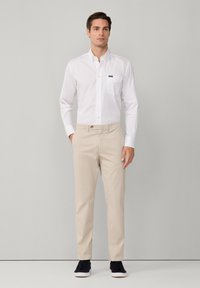 Camicia bianca con taschino sul petto, abbinata a chinos beige. Il modello indossa sneakers nere. Tessuto di cotone liscio e vestibilità su misura.