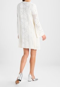 Abito lavorato a maglia bianco con maniche lunghe, caratterizzato da dettagli a occhiello e motivi floreali. Abbinato a scarpe con tacco a blocco argentate.