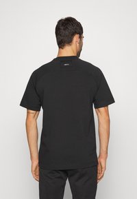 Muž v jednoduchém černém tričku s krátkým rukávem s nápisem "NIKE F.C." na horní části zad, stojící čelem k světlé šedé zdi.