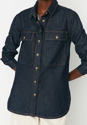 Dunkelblaues langärmliges Denimhemd mit Knopfleiste, zwei Brusttaschen und hochgekrempelten Ärmelbündchen, getragen von einer Person mit den Händen hinter dem Rücken.