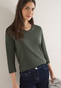 Haut vert à manches longues avec un décolleté en V, fabriqué en matière douce. Associé à un jean en denim foncé avec des surpiqûres blanches contrastantes.