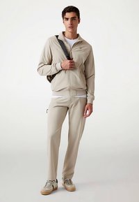 Beežist dressikomplekt lukuga jope ja pükstega, valmistatud siledast materjalist. Omadused: küljetaskud, soonikkoes mansetid ja sportlikud tennised.