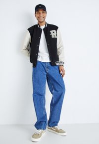 Giacca varsity nera e crema con polsini a righe, t-shirt bianca, jeans blu larghi e sneakers grigie con dettagli bianchi. Abbigliamento casual.