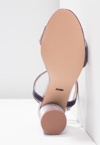 Sandale à talons hauts violette avec un design épuré, un tissu texturé et une semelle en caoutchouc beige lisse. Dispose d'une sangle à la cheville pour un meilleur maintien.