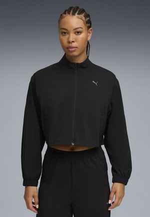 Veste de sport noire coupée en tissu léger, avec un col montant, une fermeture éclair et des poignets élastiques, arborant un petit logo sur la poitrine.
