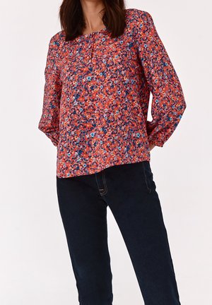 Blouse rouge à fleurs avec des manches longues, un col rond et des plis subtils au niveau de la poitrine. Jean foncé avec une silhouette ajustée.