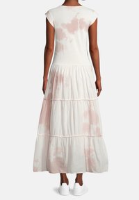 Žena v bezrukávových maxi šatech s vrstveným designem v světle růžové a bílé tie-dye barvě, obuta v bílých teniskách, zobrazená zezadu na jednobarevném pozadí.