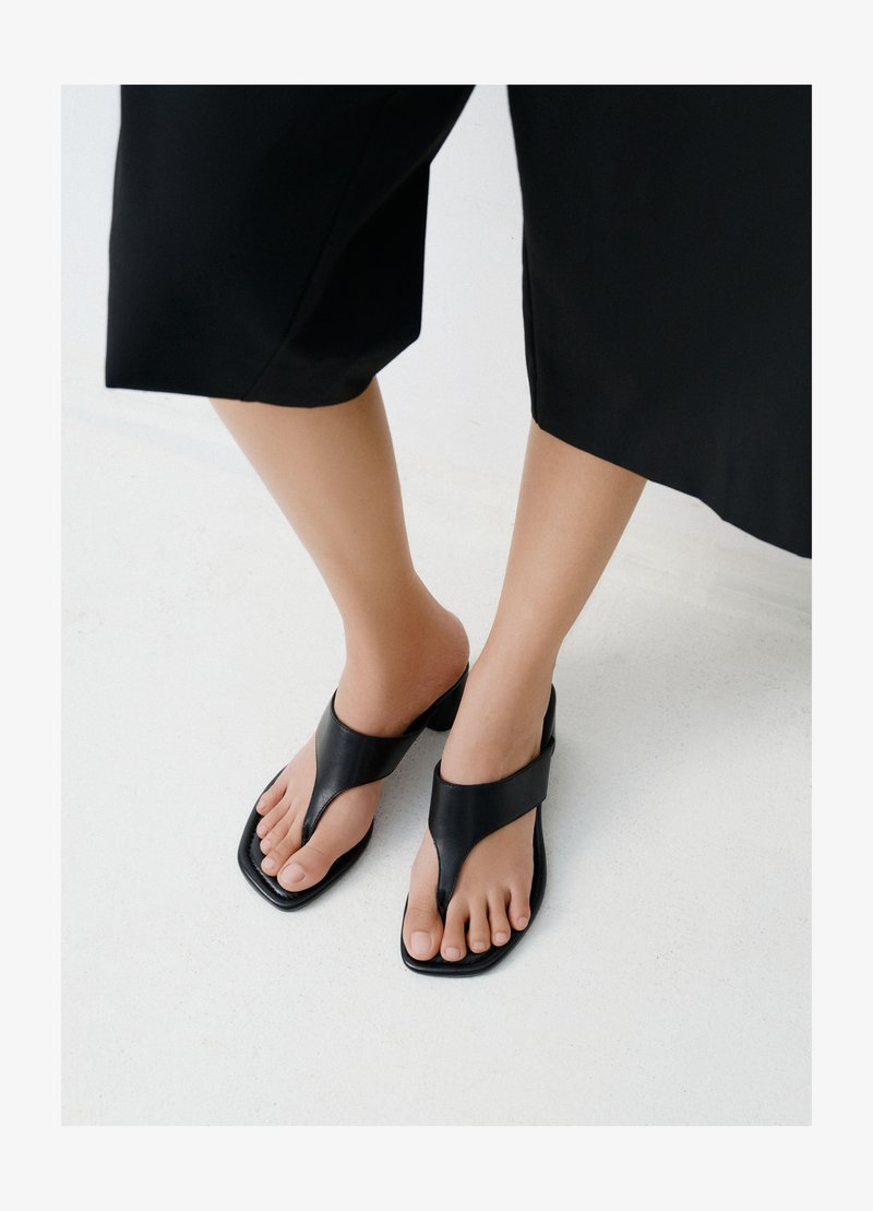 Füße, die schwarze Sandalen mit quadratischer Spitze und breiten Riemen tragen, kombiniert mit kurzen schwarzen Hosen auf einem hellen Boden.