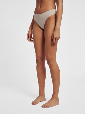 Kobieta stoi boso, ubrana w beżowe majtki bikini z wysokim wycięciem na jednolitym jasnym tle.