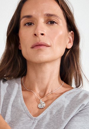 Mujer con cabello largo y castaño que lleva una camiseta gris con cuello en V y un collar plateado con un colgante abstracto, mirando hacia adelante contra un fondo liso.