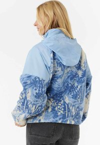 Vrouw met blond haar in een blauw gestippeld fleecejack met een effen lichtblauwe capuchon en mouwen, in combinatie met zwarte jeans, zijwaarts gekeerd.