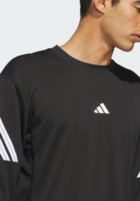 Čierne atletické tričko s dlhým rukávom má okrúhly výstrih, biely dizajn s tromi pruhmi na rukávoch a malú bielu logo na hrudi.