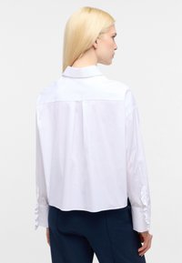 Camicia bianca corta con plissé sul retro, colletto alla mandarino e polsini arricciati. Tessuto liscio con texture minima.