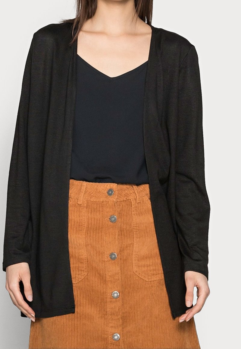 Cardigan noir à manches longues en tissu léger, porté sur un haut foncé et une jupe en velours côtelé orange avec des boutons sur le devant.