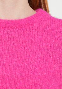 Gaiši rozā mīksti adīts džemperis ar ribotu apaļu apkakli; tam ir maiga tekstūra un nelielas krāsas nianses visā virsmā.