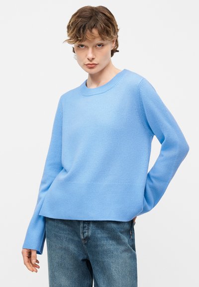 Νεαρό άτομο που φοράει ανοιχτό μπλε πουλόβερ και μπλε τζιν, στέκεται με το ένα χέρι πίσω από την πλάτη, μπροστά σε λευκό φόντο.