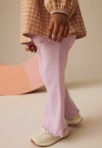 Rosa ribbade byxor med vida ben och scallopdetalj i nederkant, kombinerat med ljusa sneakers. Överlagd med en rutigt blus i senapsgul färg. Bakgrund i mjuka pastellfärger.