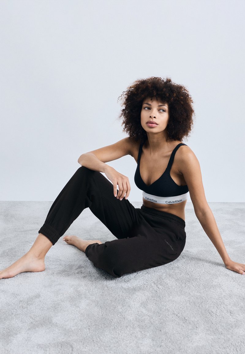 Juodas braletas su Calvin Klein logotipu, derinamas su laisvomis juodomis treniruočių kelnėmis, sėdint ant šviesiai pilkos tekstūruotos paviršiaus.