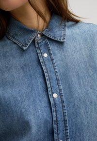 Denim shirt with a classic collar, featuring silver snap buttons and contrasting orange stitching on a medium blue fabric.