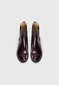 Une paire de bottes Chelsea en cuir brun foncé poli avec des panneaux latéraux élastiques noirs et une doublure intérieure beige sur fond blanc.