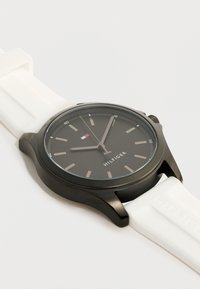 Μαύρο στρογγυλό καντράν ρολογιού με μπεζ δείκτες ώρας, ασημένια δείκτες, μικρή κόκκινη, λευκή και μπλε σημαία, και λουράκι λευκής σιλικόνης σε λευκό φόντο.