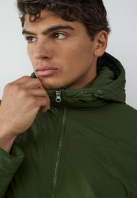 Giacca leggera verde con chiusura a zip e cappuccio regolabile; presenta una consistenza liscia e una leggera lucentezza. Mano che afferra il colletto.