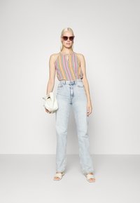 Top de halter colorido com padrão, jeans de cintura alta azul claro, saltos abertos de cor bege clara, segurando uma bolsa branca, com óculos de sol rosa.