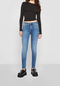 Mujer vestida con jeans ajustados azules, un top negro de manga larga y cropped, y zapatos de plataforma negros, de pie frente a un fondo gris claro liso.