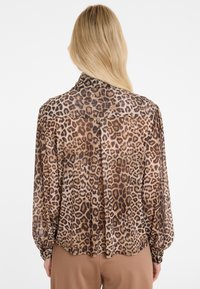 Woman with long blonde hair wearing a loose, long-sleeve leopard print blouse and brown pants, shown from the back against a white background.