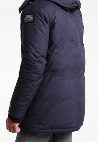 Marinblå vadderad jacka med huva, med en logotyppatch på axeln och elastiska ärmslut. Slät textur och skräddarsydd passform.