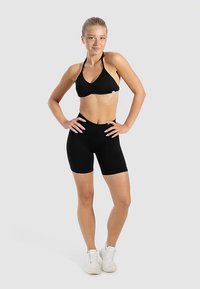 Svart halterneck-bikinitopp med rynkad design och matchande höga shorts. Modellen bär vita träningsskor. Enfärgad grå bakgrund.