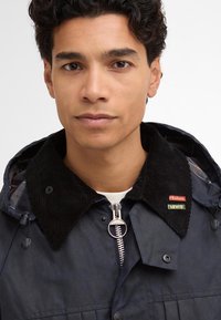 Casaco capucho azul-marinho com colarinho de veludo corduroy, fecho de correr prateado e pins de marca da Barbour e Levi's. Possui um exterior texturizado e forro xadrez.