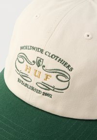 Beżowa i zielona czapka z daszkiem z wyhaftowanym napisem "WORLDWIDE CLOTHIERS HUF ESTABLISHED 2002" w zielonym i żółtym kolorze nici na przednim panelu.