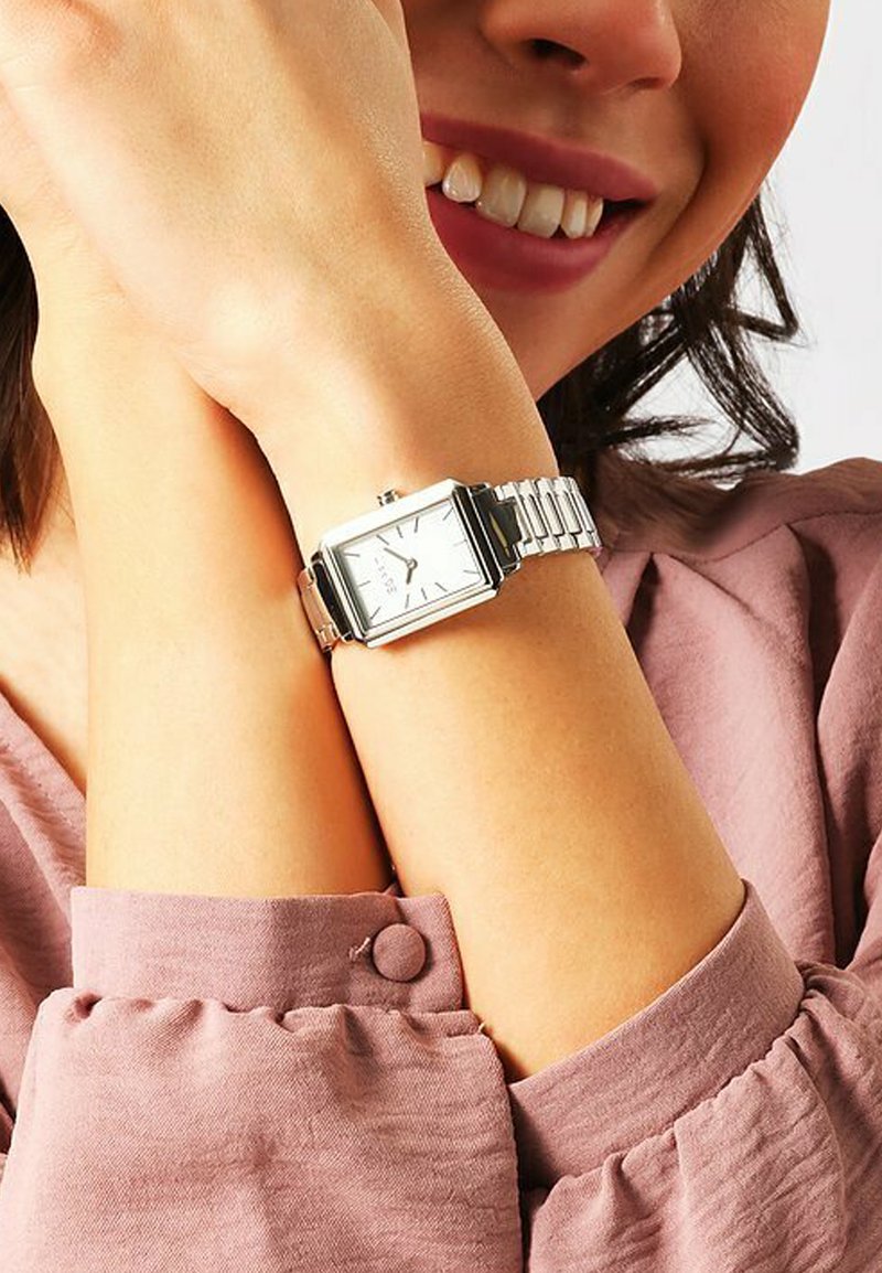 Silberne rechteckige Uhr mit weißem Zifferblatt, Metallarmband und minimalistischem Design. Trägt am Handgelenk mit einem hellrosa Stoffhemd.