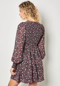 Czarna sukienka w kwiaty z marszczoną talią, długimi bufkami i rozszerzonym dołem, ozdobiona różowymi i białymi wzorami kwiatów na przezroczystym materiale.