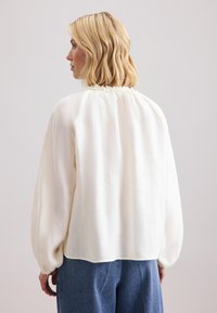 Blusa bianca leggera con scollatura arricciata e maniche lunghe. Tessuto morbido con vestibilità rilassata, abbinata a pantaloni di jeans blu.