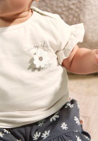 Hellbeiges kurzärmeliges Hemd mit Rüschen an den Kanten, versehen mit einer Blumenapplikation und gesticktem Text; kombiniert mit marineblauen, floral gemusterten Hosen.