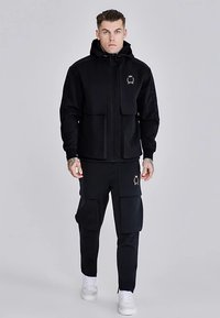 Čierna fleecová mikina na zips s dvoma prednými vreckami a zlatým logom, v kombinácii s nohavicami typu cargo s veľkými bočnými vreckami a bielymi teniskami.