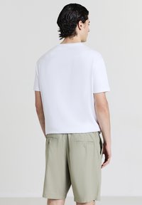 Homme aux cheveux foncés portant une chemise blanche à manches courtes simple et un short beige clair, se tenant de dos contre un fond clair.