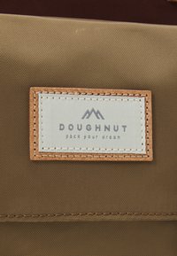 Brūna auduma birka ar tekstu "DOUGHNUT" tumši pelēkā krāsā un kalnu grafiku. Adīts ādas mala piešķir tekstūru.