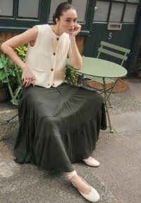 Light beige, textured sleeveless vest with front buttons paired with a long, dark green, pleated skirt and cream-colored ballet flats.