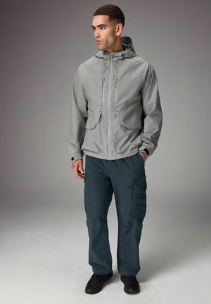 Helle graue wasserdichte Jacke mit Kapuze und zwei Fronttaschen, kombiniert mit dunkelblauen Cargo-Hosen mit Seitentaschen. Schwarze Sneaker runden das Outfit ab.