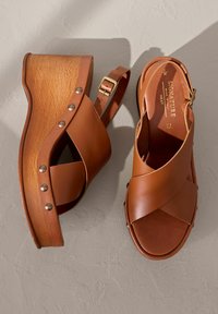 Sandali wedge in pelle marrone con cinturini incrociati, tacco in legno, borchie metalliche e cinturino alla caviglia regolabile. La texture è liscia.