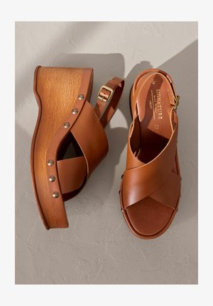 Braune Leder-Plateausandalen mit überkreuzten Riemen, Holzabsatz, metallischen Nieten und verstellbarem Knöchelriemen. Die Textur ist glatt.