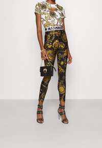 Weißes kurzärmeliges Top mit Ausschnitten, gemustert mit goldenen Designs, kombiniert mit schwarzen Leggings mit goldenen Akzenten. Schwarze Handtasche und mit Nieten besetzte High Heels.