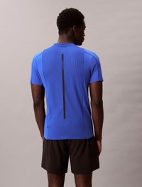 Camisa atlética azul de mangas curtas, com textura em malha e um design de faixa preta vertical nas costas, combinada com calções pretos.