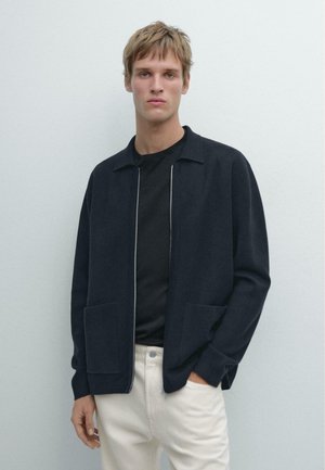 Giacca blu navy con zip realizzata in materiale morbido, con colletto, due tasche frontali e vestibilità comoda, abbinata a una camicia nera e pantaloni chiari.